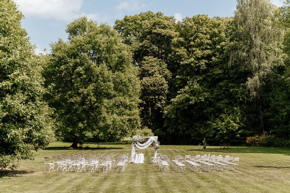A panoramic view of an outdoor wedding ceremony setup with white chairs arranged in rows on a lush green lawn surrounded by trees.