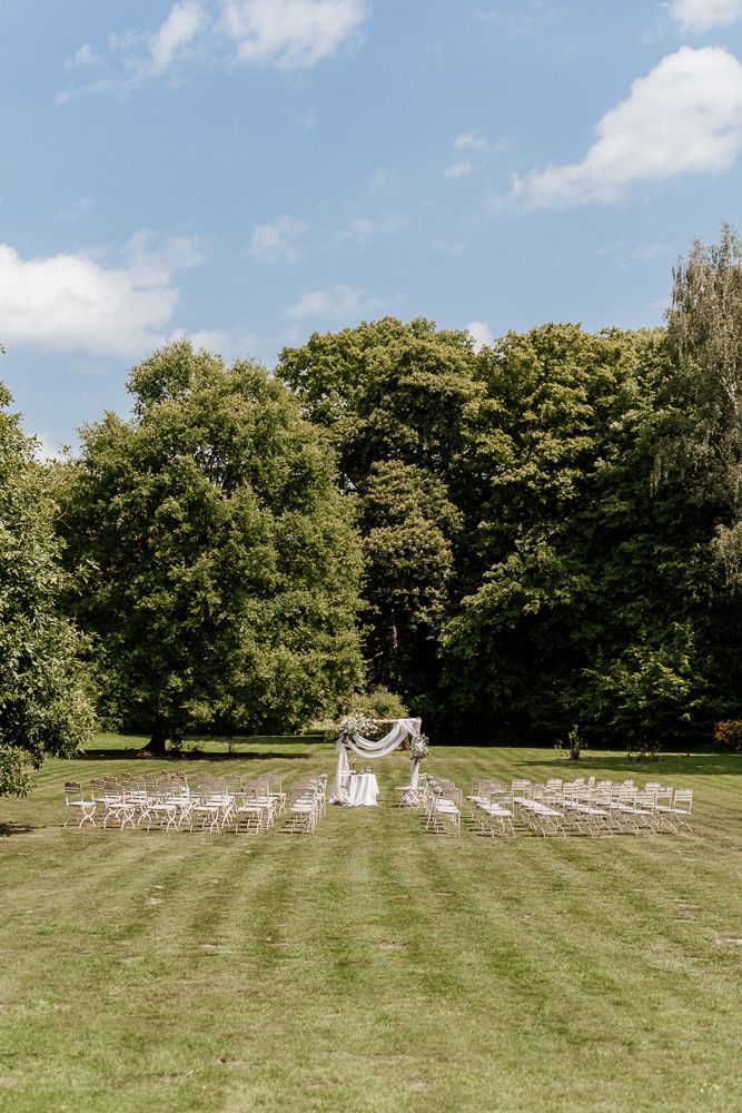 Outdoor wedding ceremony setup with white chairs arranged on a green lawn surrounded by trees.