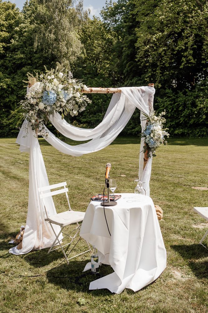 White fabric wedding arch with floral arrangements stands elegantly in a sunny garden setting.