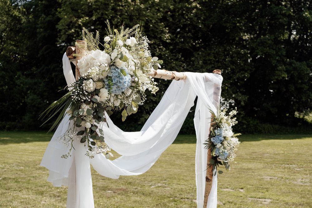 Elegant outdoor wedding setup featuring draped white fabric arch adorned with white floral arrangements.