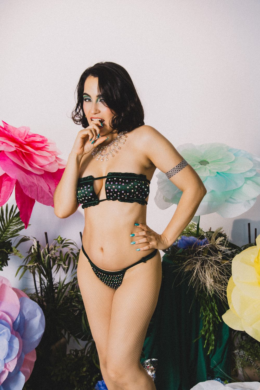 Woman in black polka dot bikini posing among colorful paper flowers and tropical plants in a studio setting.