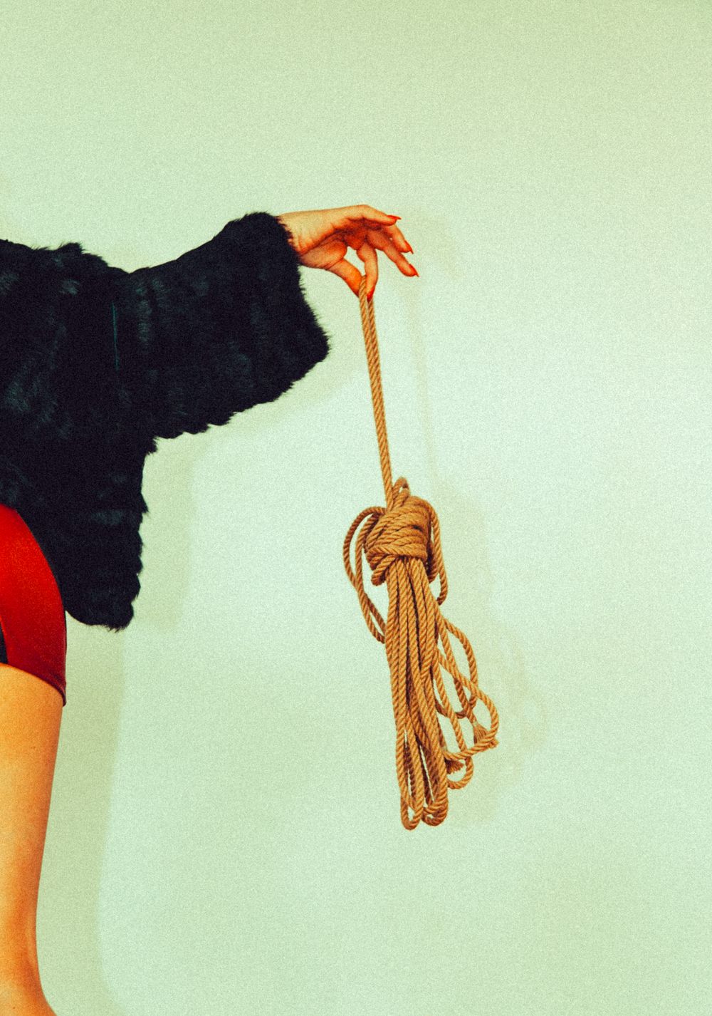 Woman in black fur coat holding coiled natural rope against mint green background.