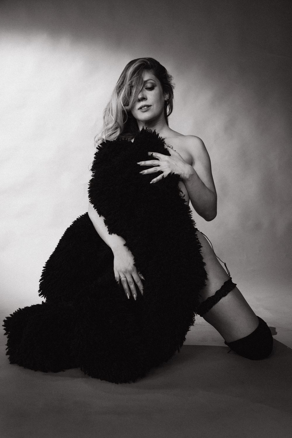 Black and white studio photo of a blonde woman posing elegantly in a fluffy black coat, seated on the floor.