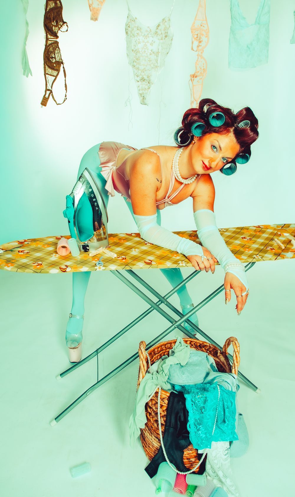 Retro pin-up woman with hair rollers ironing clothes near a laundry basket in a teal-themed studio setup.
