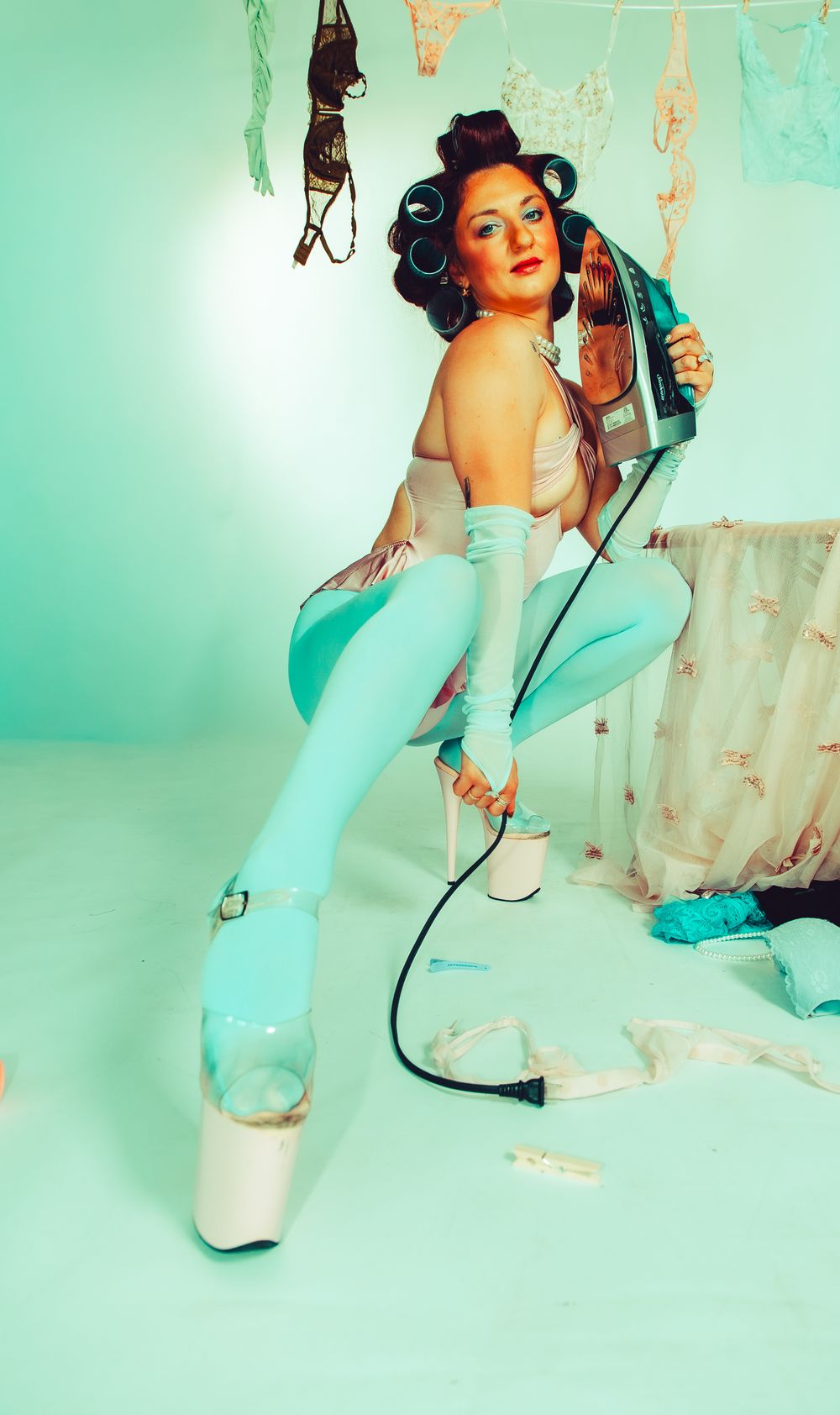 Woman in blue leggings and platform heels holds an iron, styled with hair rollers in a retro pin-up themed photo shoot.