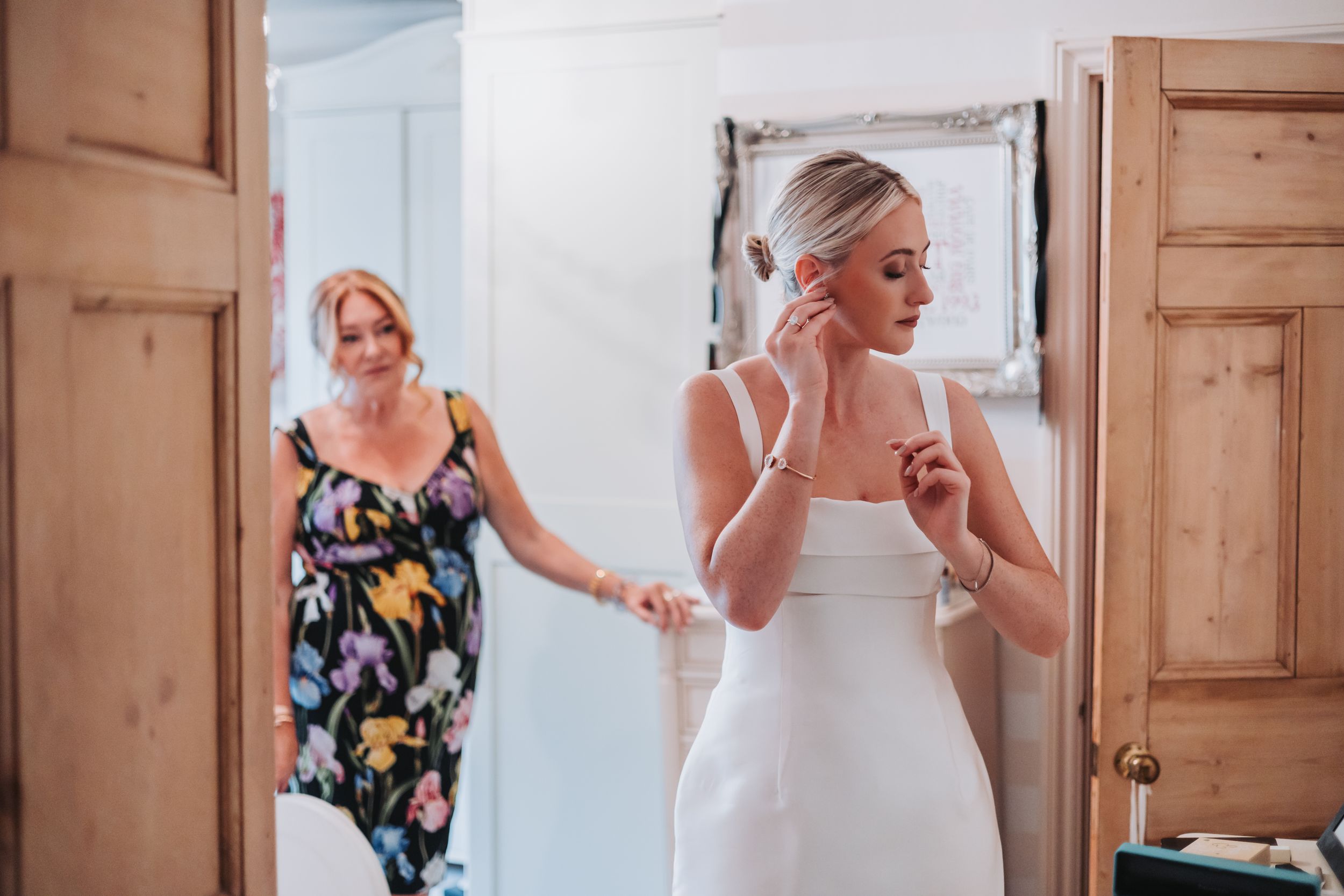 Bride in white dress adjusting earring while woman in floral dress assists during wedding preparations.