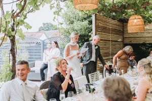 Bride and groom giving speech at outdoor wedding reception with guests seated at decorated tables.