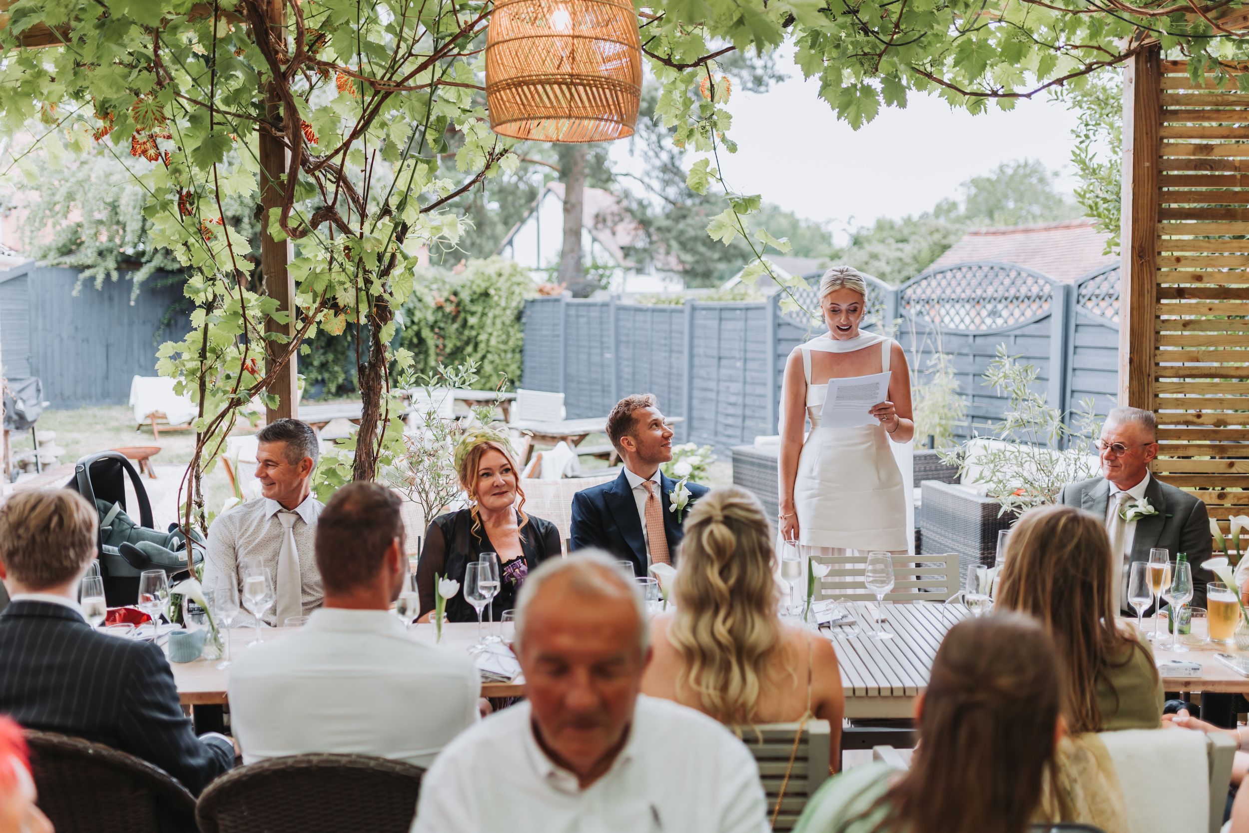Bride in white dress giving speech at outdoor wedding reception with guests seated at decorated tables under trees.