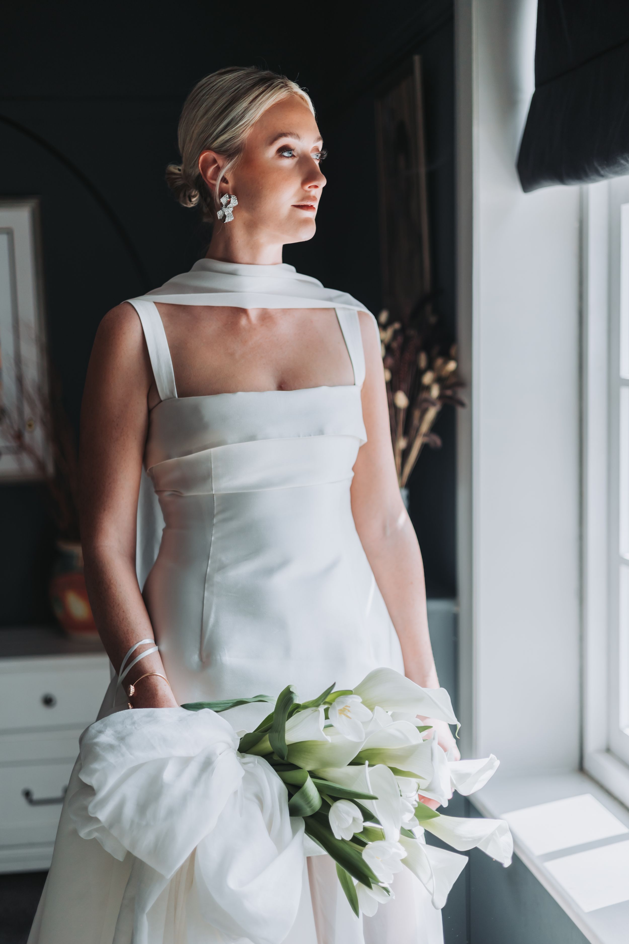 Elegant bride in white gown holding tulip bouquet stands by window in soft natural light.