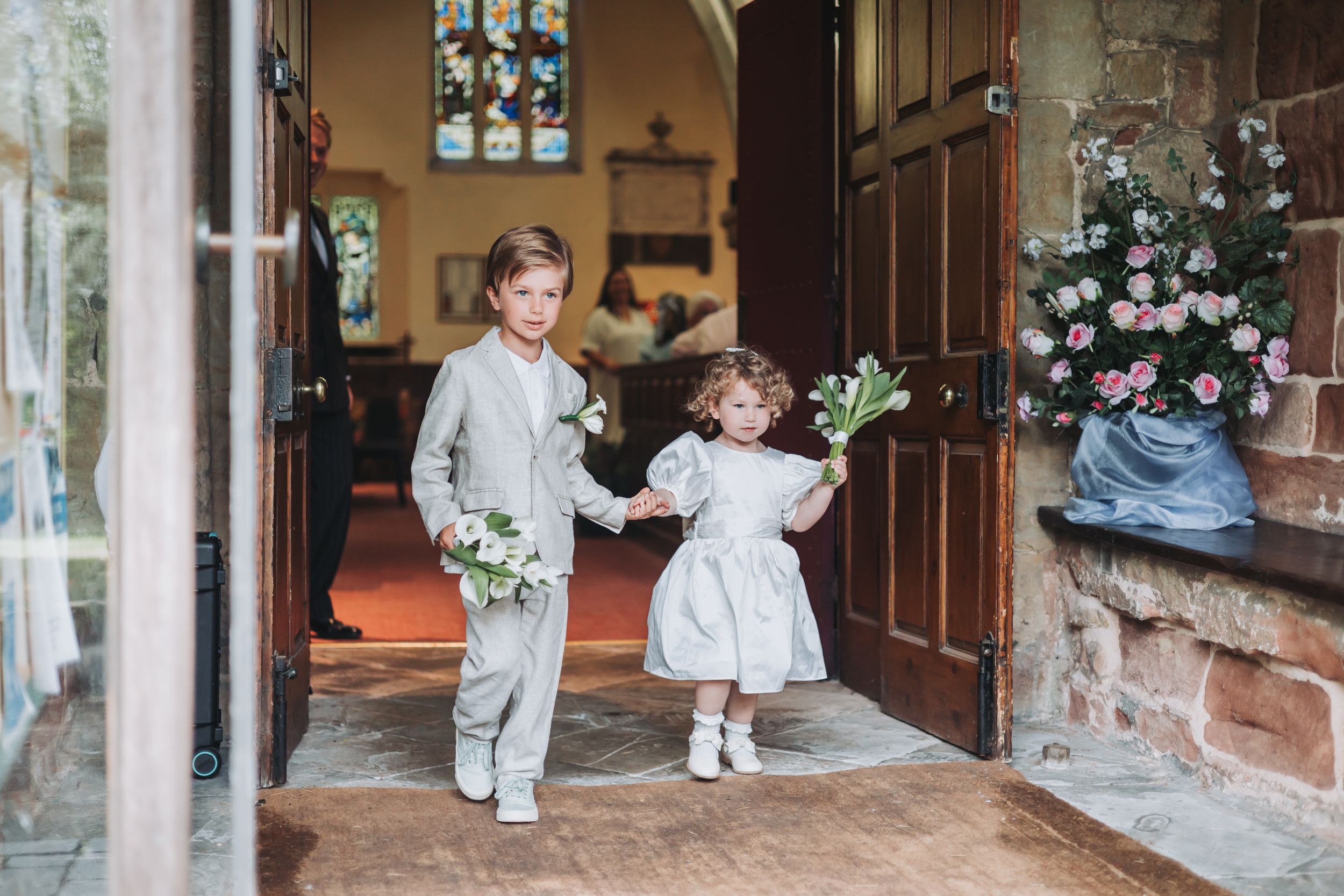 Two children in formal wedding attire hold flowers and walk hand in hand through a church doorway.