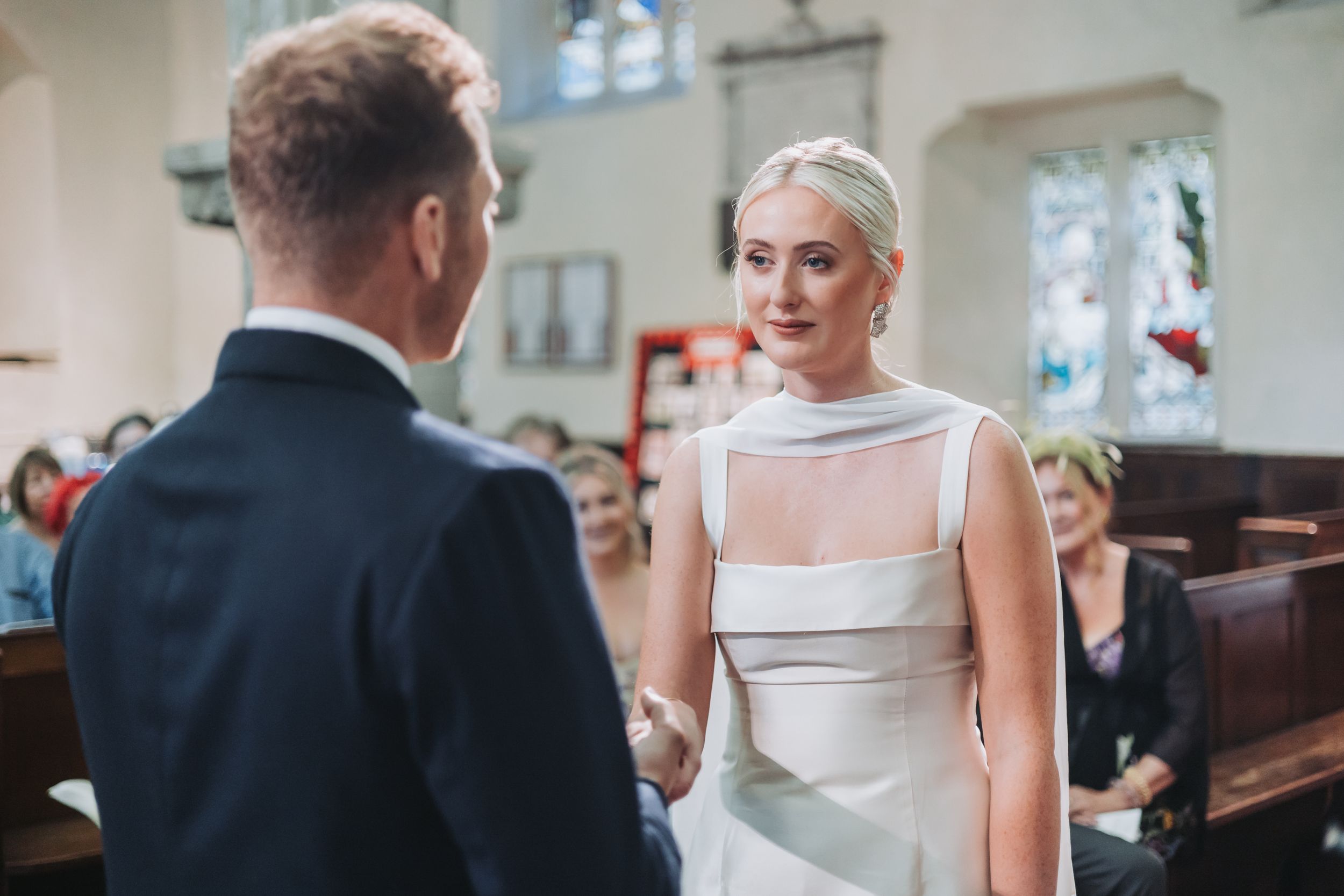 Bride in sleek white gown holds hands with groom during emotional church wedding ceremony.