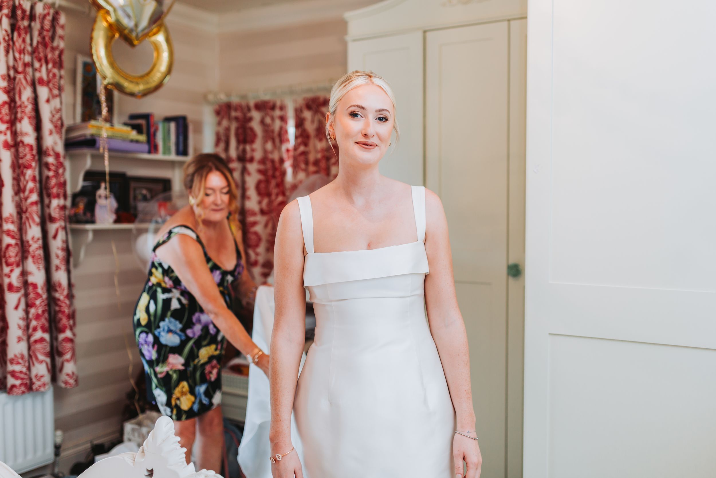 Bride in white dress getting ready with bridesmaid helping in floral-decorated room with gold balloon.