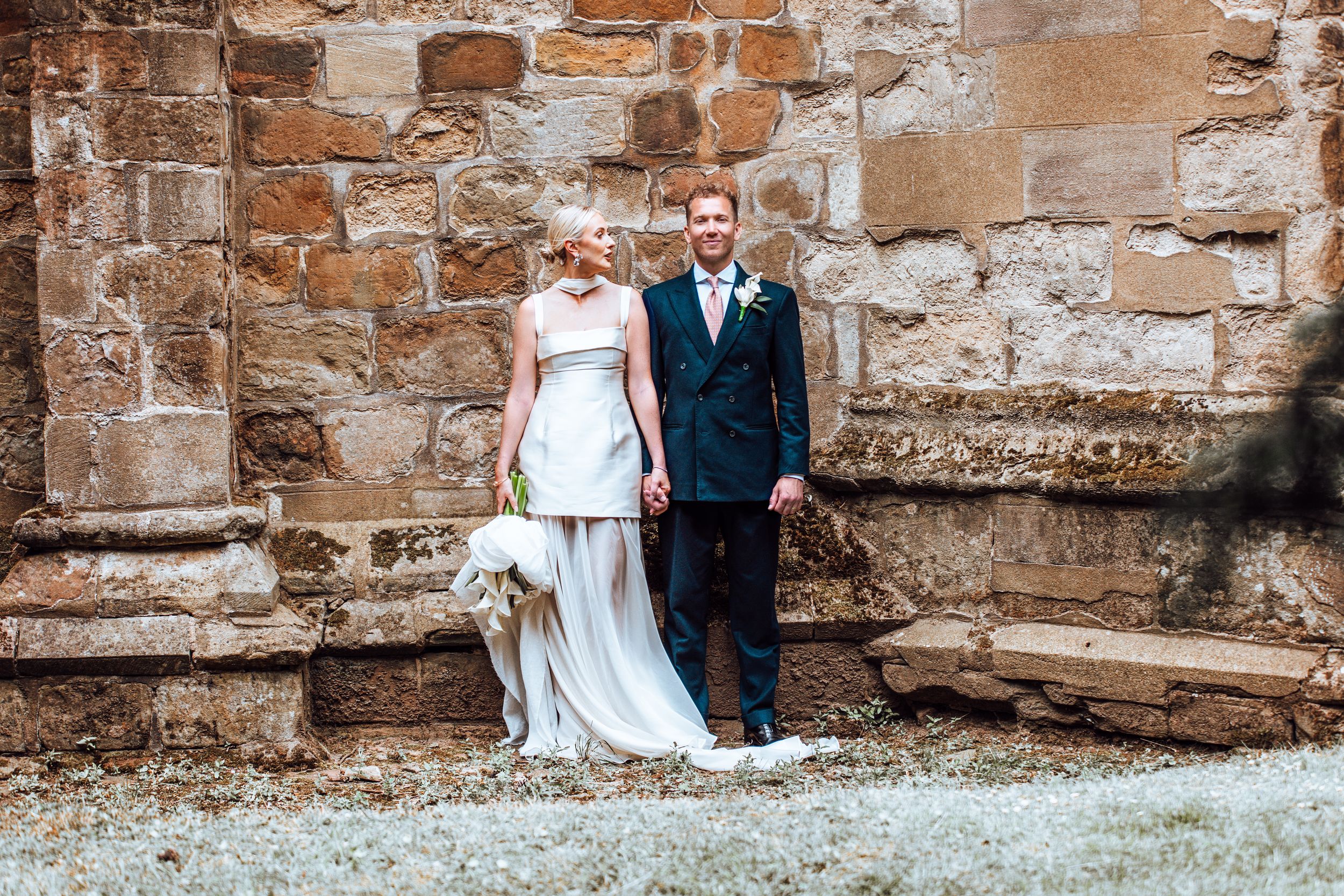 A wedding couple stands against a rustic stone wall at an ancient abbey or castle ruins.