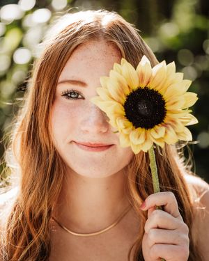 Red head senior girl holding a sunflower over one of her blue eyes during her senior photo session.
