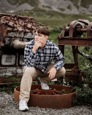 Senior boy plaid shirt and khakis sits thoughtfully on rustic metal equipment in an outdoor setting.