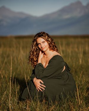 Curly haired senior girl wearing green dress crouching down in field in front of mountain landscape at golden hour during her senior session