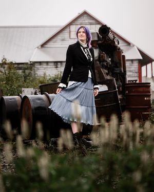 Senior girl with purple hair in a pleated skirt and dark top poses against a rustic barn setting in moody outdoor lighting.