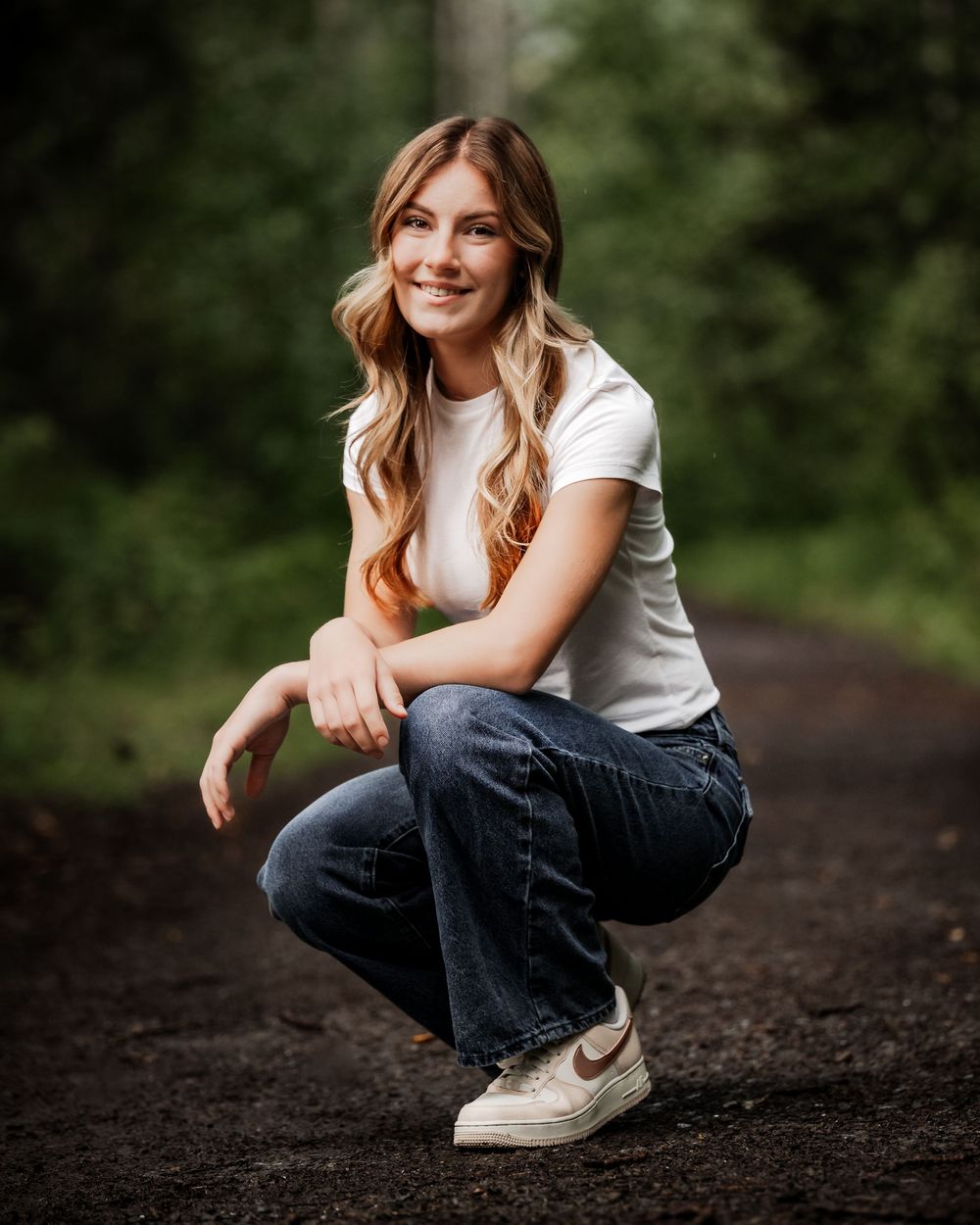 Senior girl white shirt, jeans and Nike sneakers crouches down on in outdoor setting with blurred green foliage background.