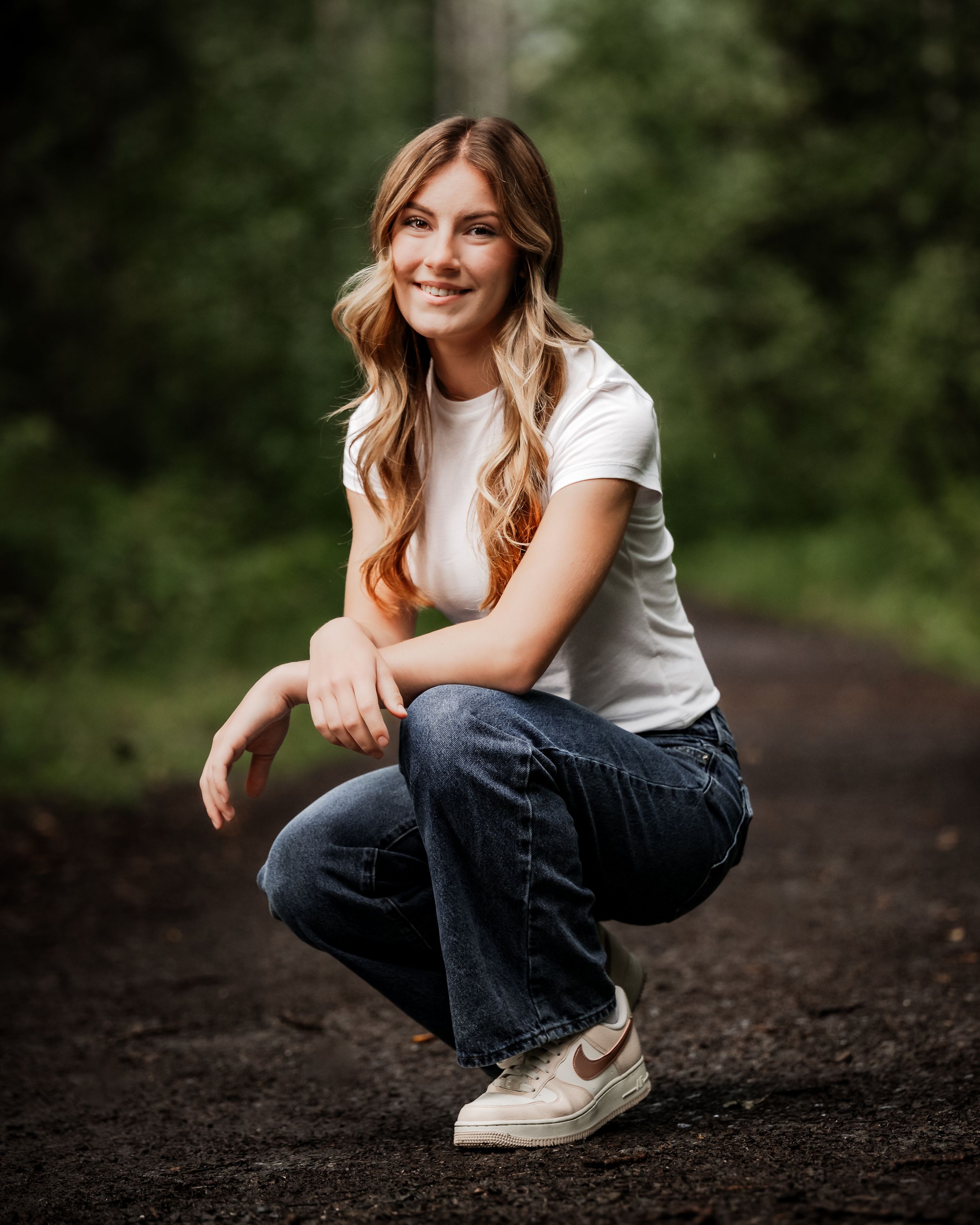 Senior girl white shirt, jeans and Nike sneakers crouches down on in outdoor setting with blurred green foliage background.