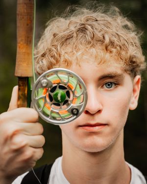 Blonde curly hair boy holding a fly fishing rod over one of his eyes during his senior photo session.