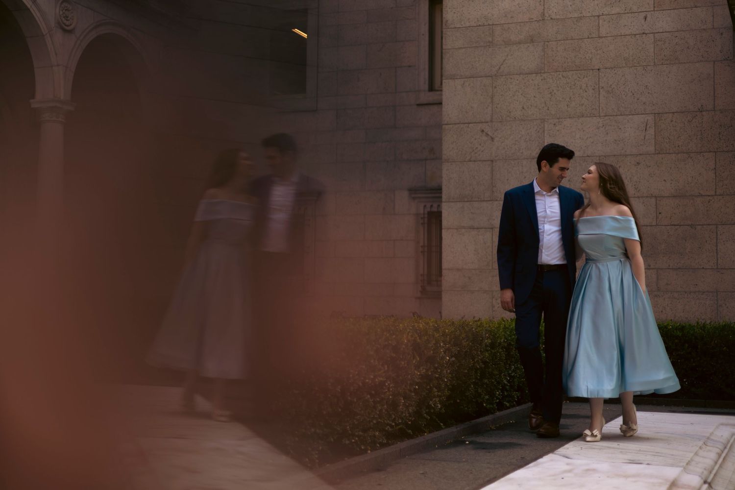 Two figures in formal attire walk together in an elegant setting with warm lighting and stone walls.
