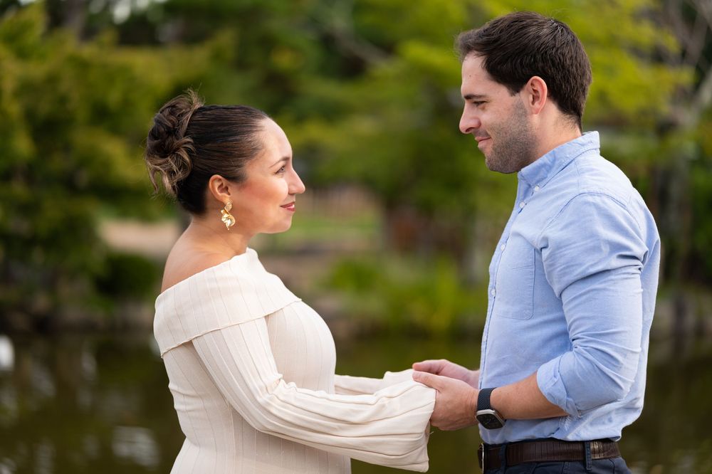 Expecting couple holding hands and smiling at each other beside a serene Japanese garden pond.