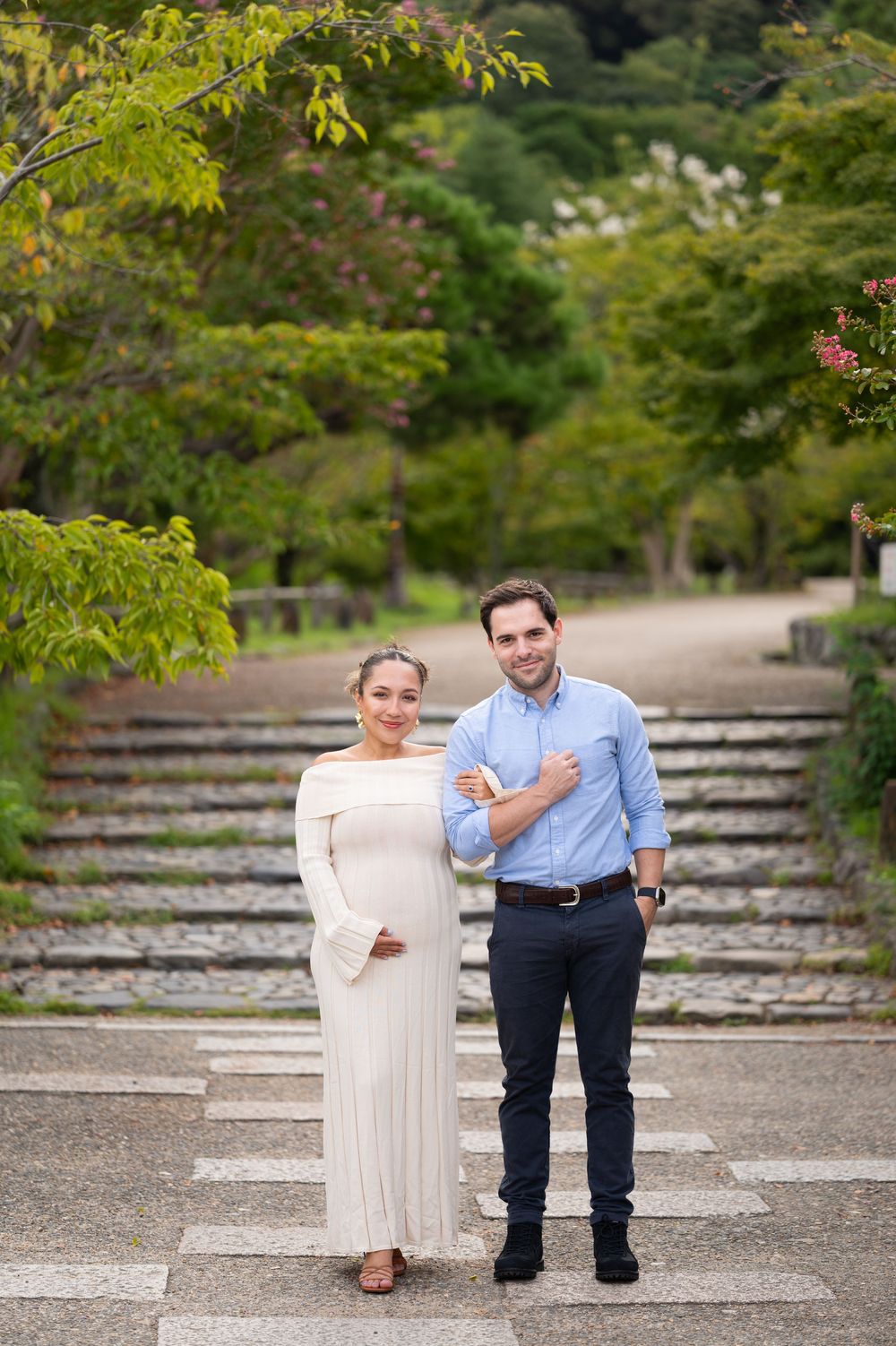 Smiling pregnant couple posing together on stone steps in a lush green Japanese garden.