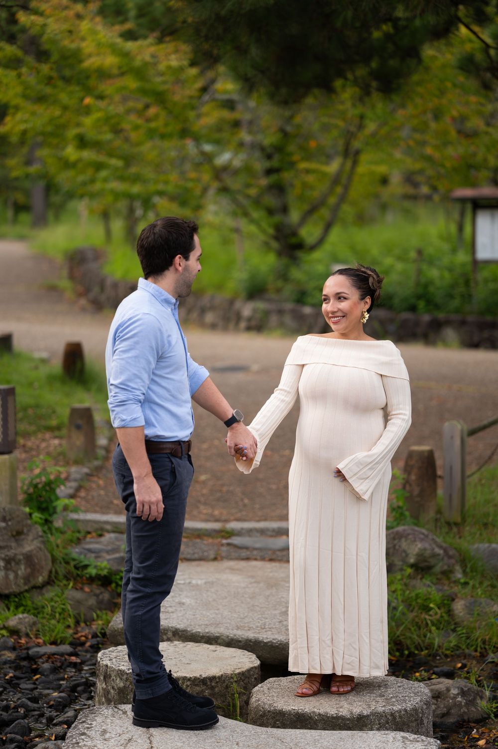Pregnant couple holds hands on stepping stones in a lush Japanese garden, woman in white dress.