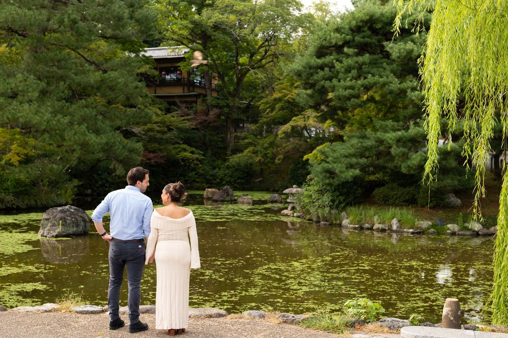 Pregnant couple standing together admiring a tranquil Japanese garden pond with a willow tree.