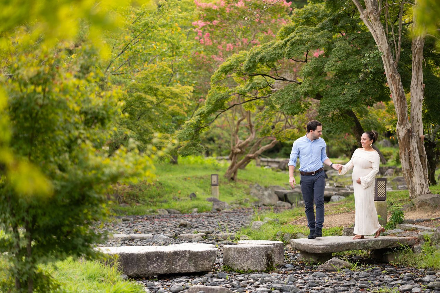 Couple walks hand-in-hand across stepping stones in a serene Japanese garden with green foliage.