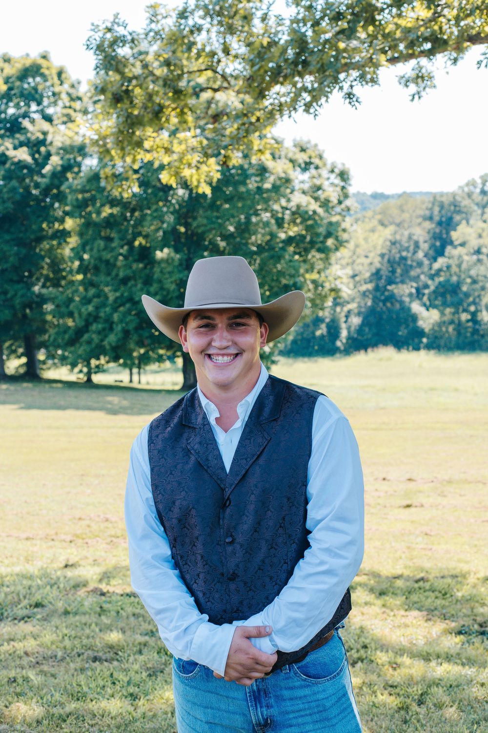 A western-styled groomsman in denim, vest and cowboy hat poses in a sunny field.