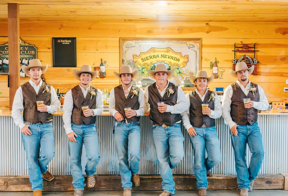 Groomsmen in matching denim and vests lean against a rustic wooden bar.