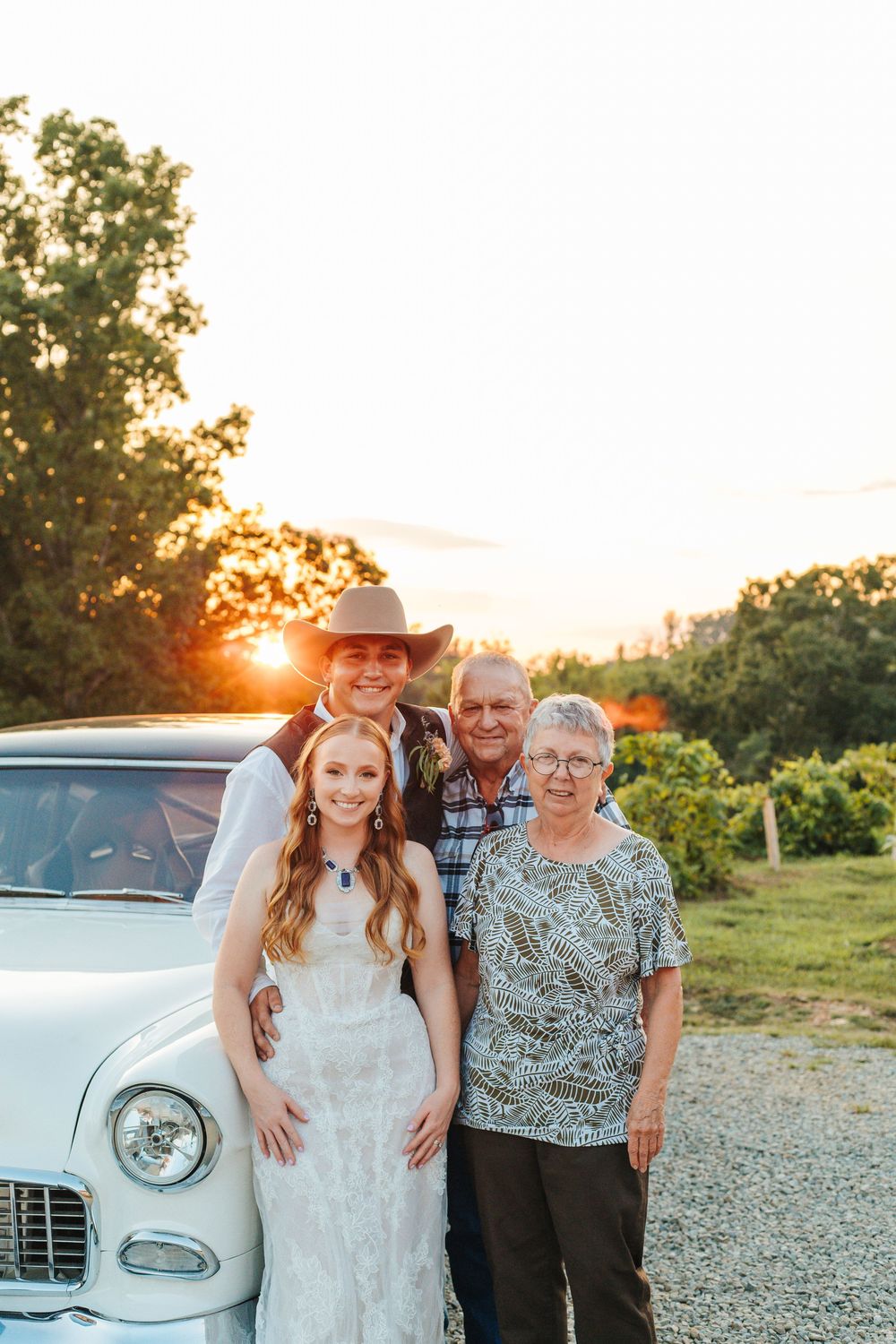 A wedding party poses together beside a vintage car at sunset.