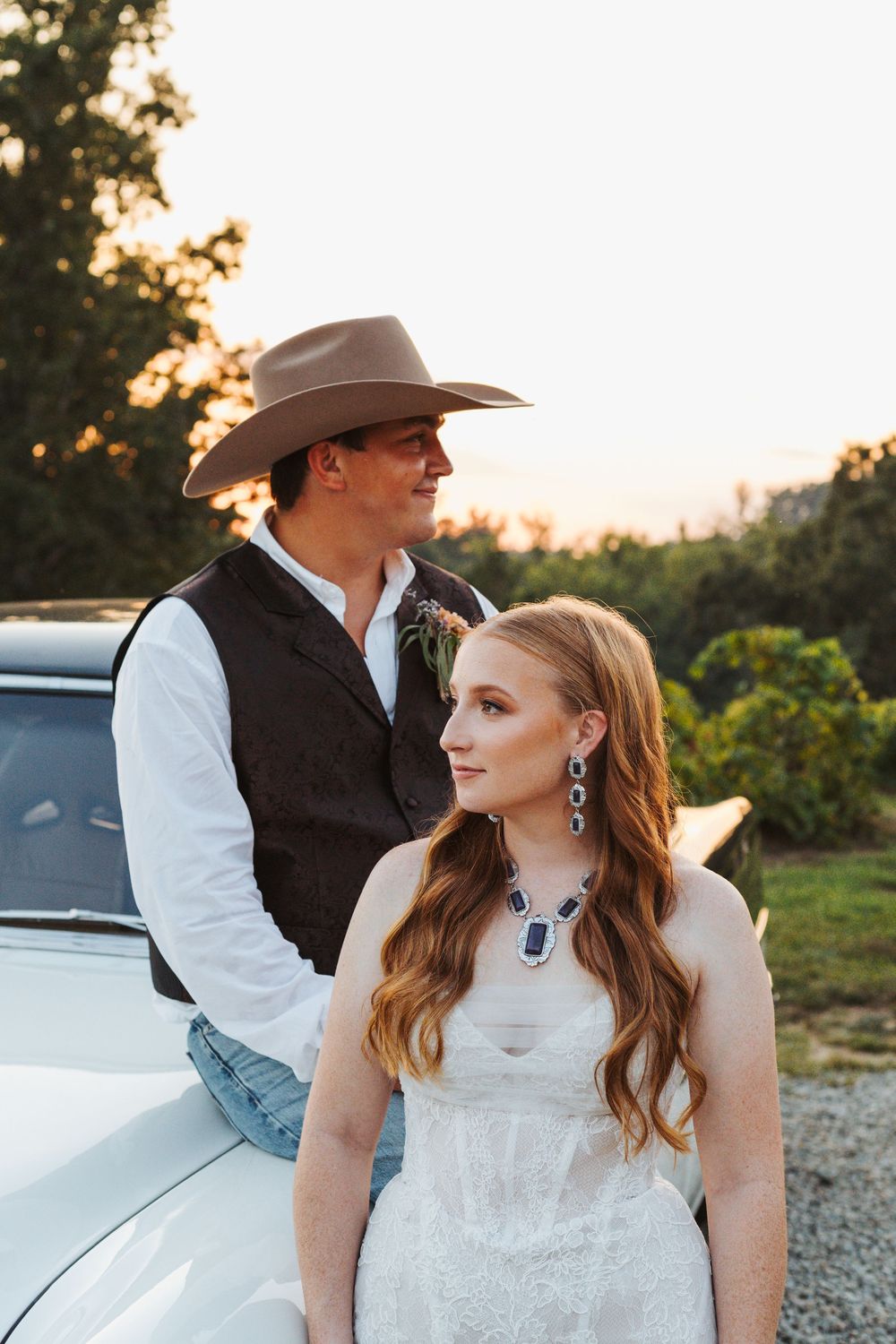 A couple in western wedding attire pose against a classic car during golden hour.