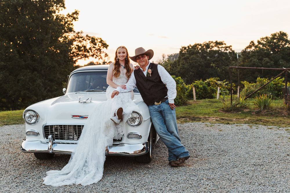 Newlyweds pose with a vintage white Chevrolet at their rustic countryside wedding.
