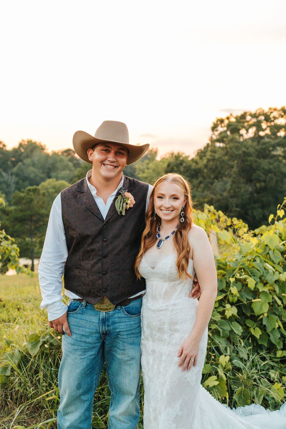 A country wedding couple in cowboy attire stands together in a vineyard at sunset.