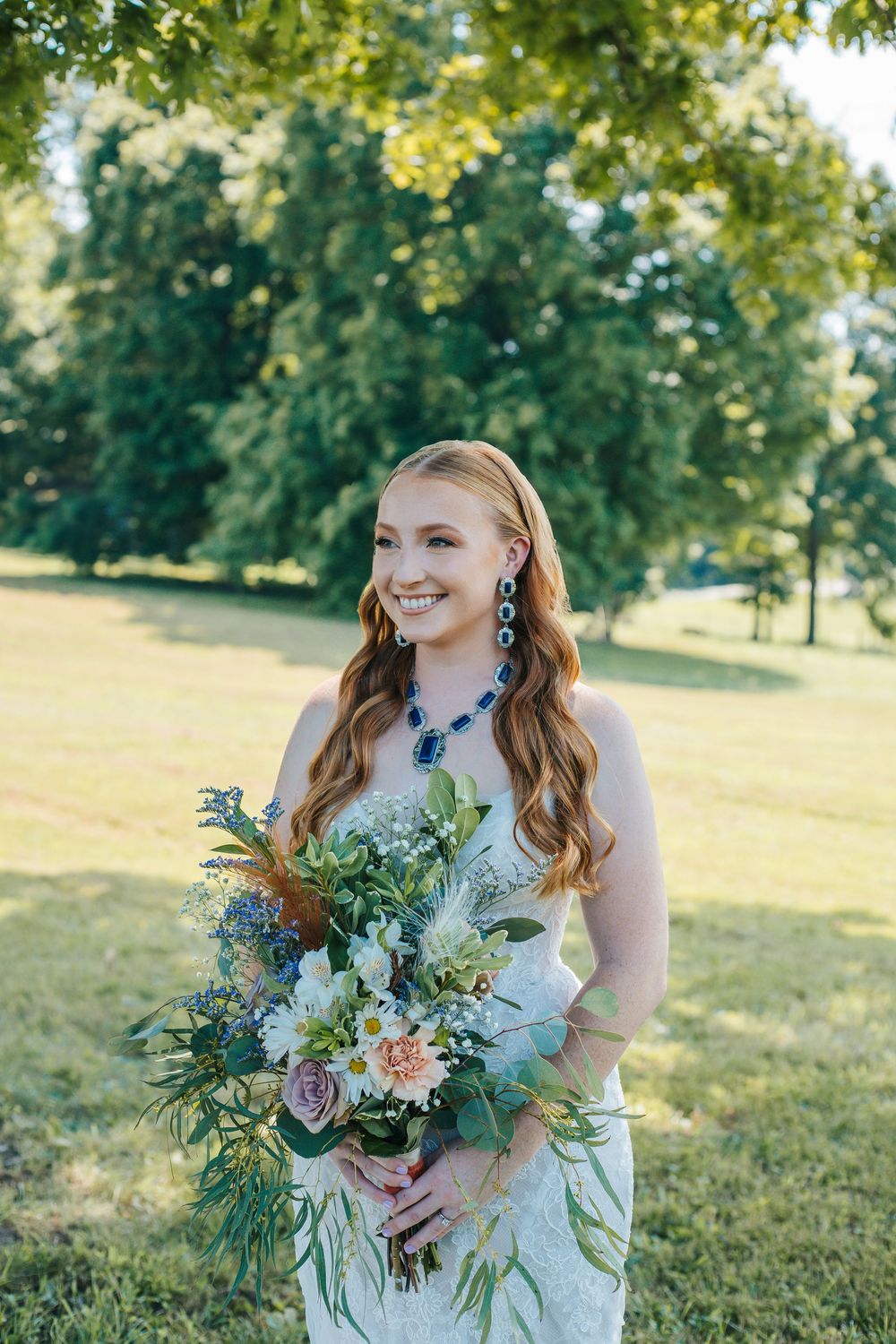 A bride in a lace wedding dress holds a stunning wildflower bouquet in an outdoor garden setting.