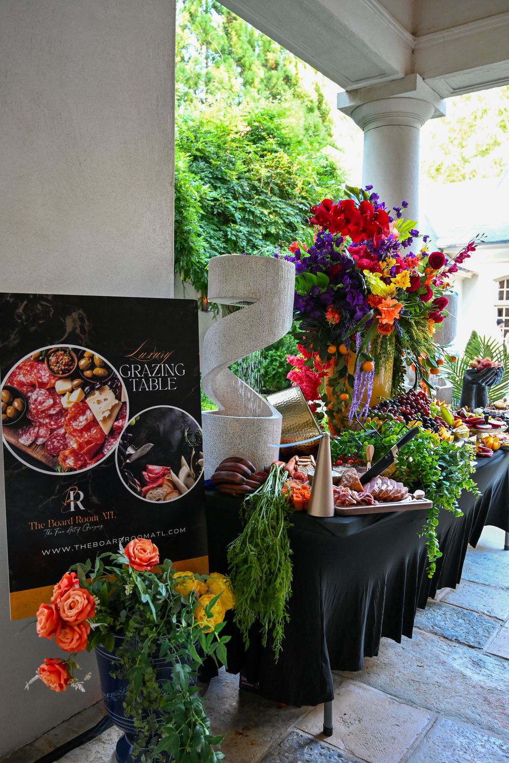 Elegant catering display features black tablecloths adorned with vibrant floral arrangements in purple, orange and red blooms.