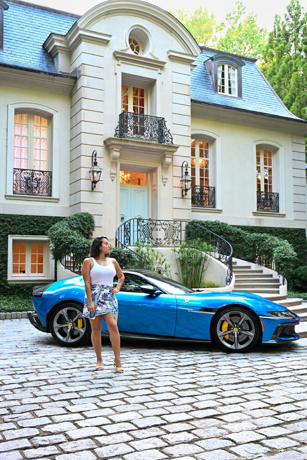 A bright blue Ferrari sports car parked in front of a luxurious French-style mansion with stone facade and cobblestone driveway.