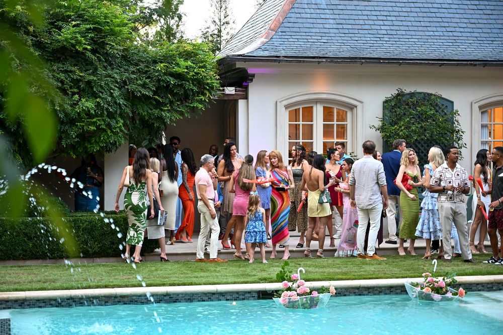 People gather for a summer party around an outdoor swimming pool with a white house visible in the background.