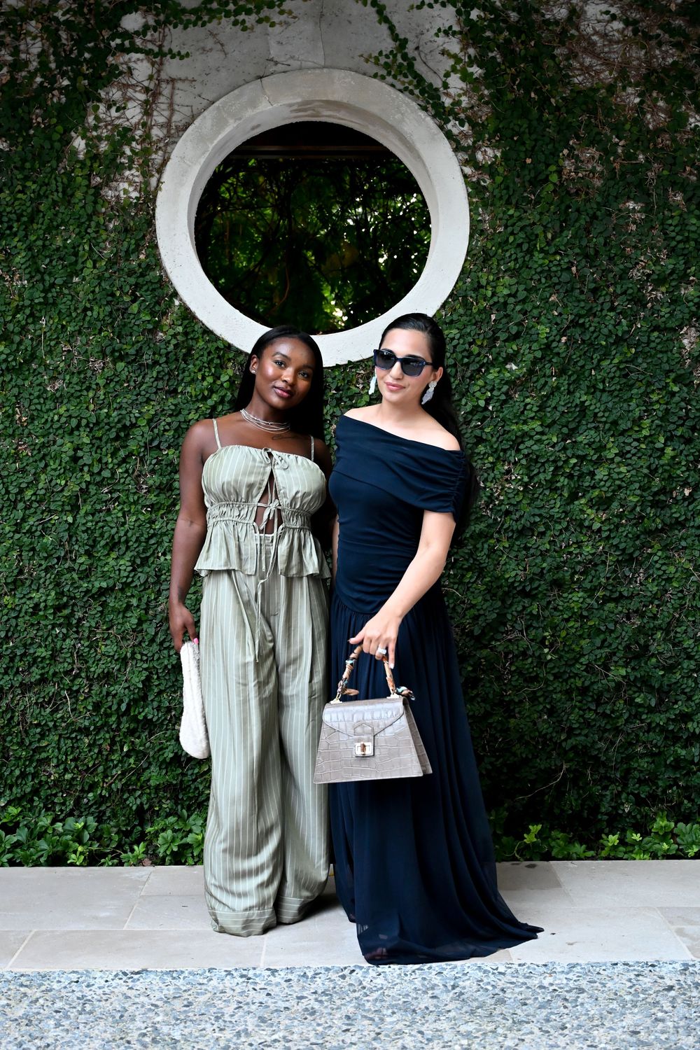 Two people sit on a white bench against a lush green hedge wall with a circular window feature.