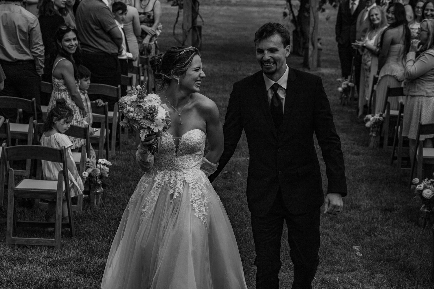 A bride and groom joyfully walk together on grass after their outdoor wedding ceremony.
