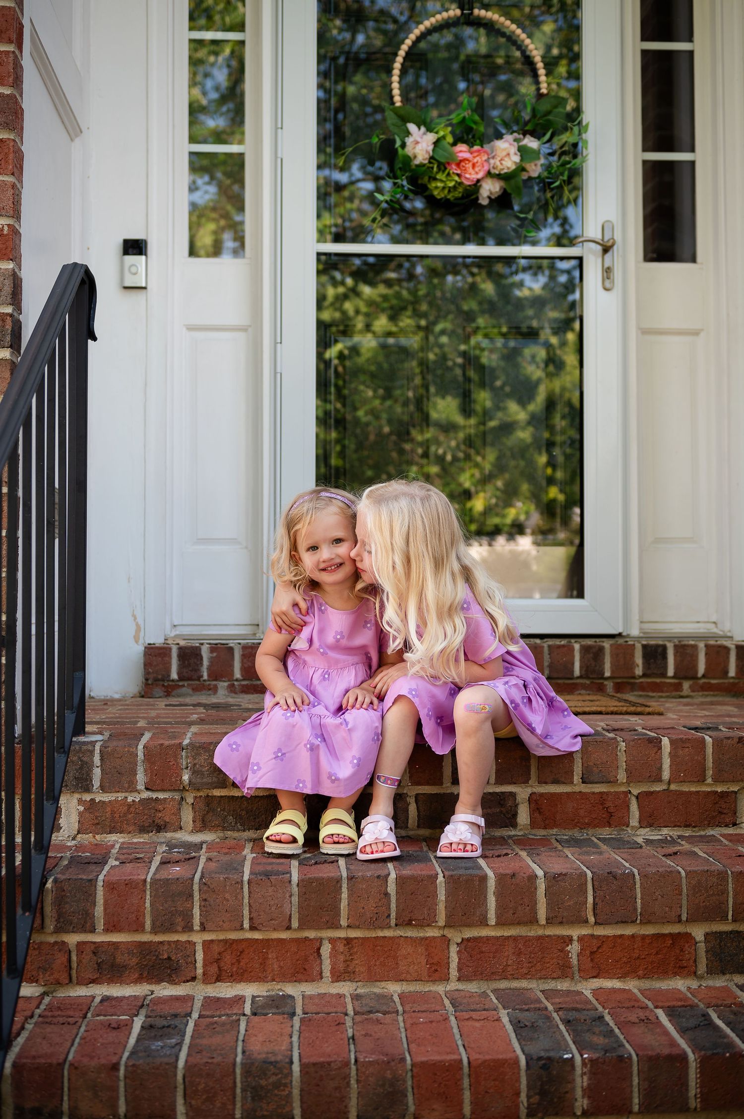 Two young children in pink dresses sitting together on brick front porch steps.