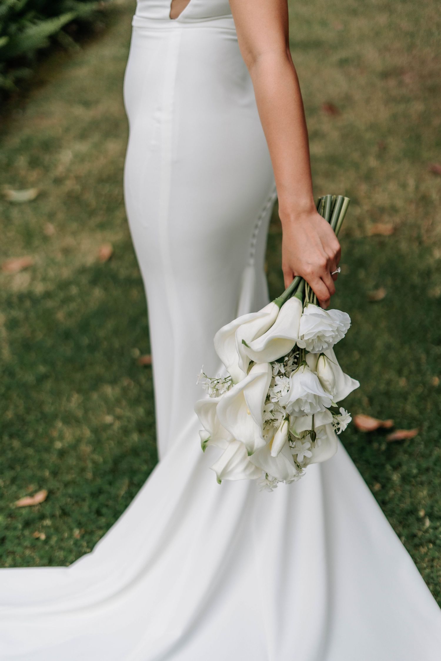 Close-up details of an elegant white bridal gown with trumpet silhouette and someone holding a white floral bouquet.