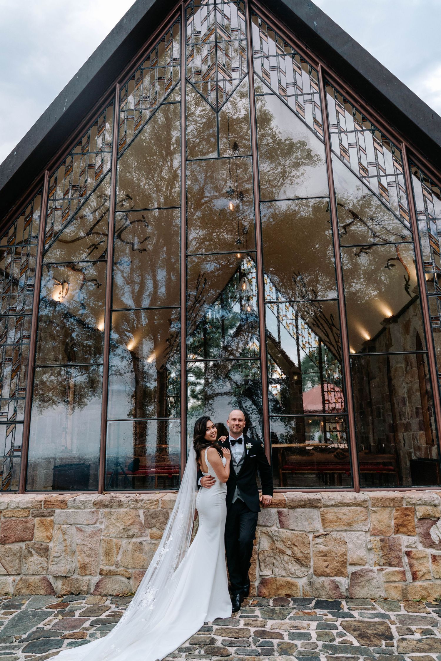 A wedding couple poses together against a stunning stone and glass greenhouse chapel exterior in a romantic series.