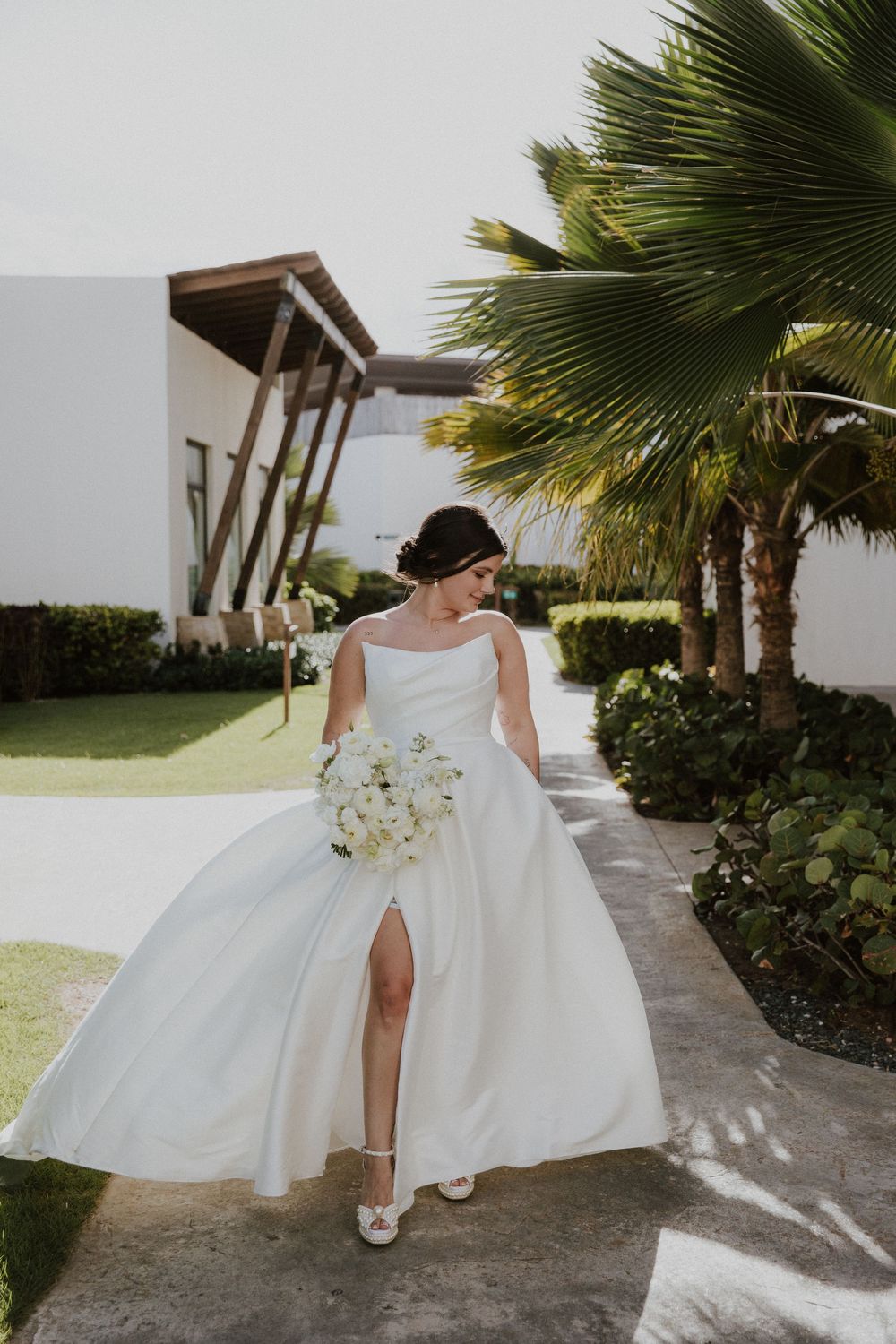 Bride in flowing white wedding dress walking along outdoor pathway with palm trees and white walls in Mexico.