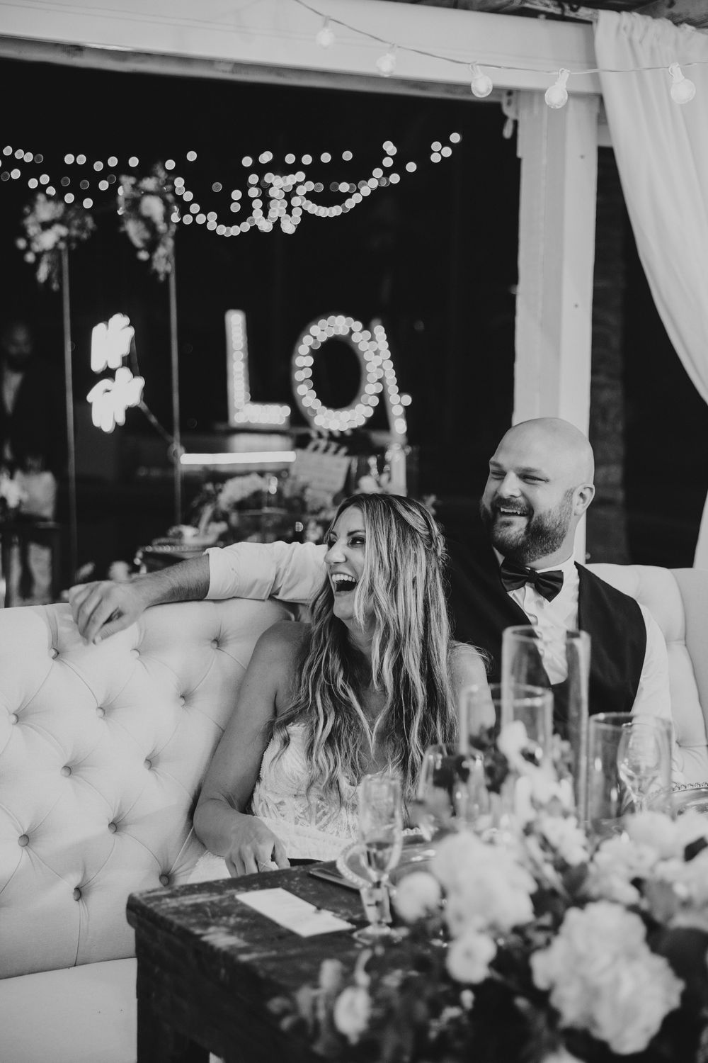 Black and white wedding reception photo with couple sitting at decorated table with floral centerpiece and twinkling lights overhead.
