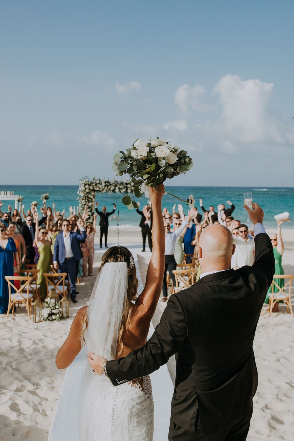 A beachside wedding ceremony with guests standing in white chairs on sandy shores against turquoise ocean waters.