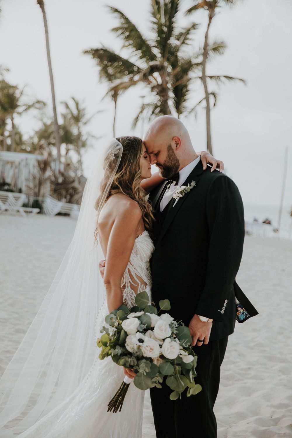 A romantic beach wedding couple embraces with white flowers and palm trees swaying in the background.