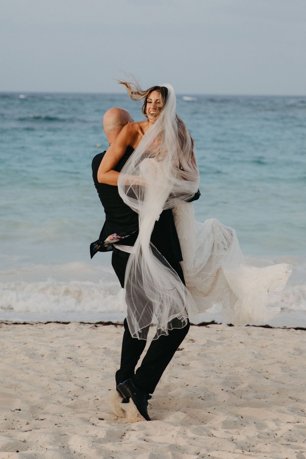 Dancing couple embraces on a tropical beach with flowing white veil caught in ocean breeze.